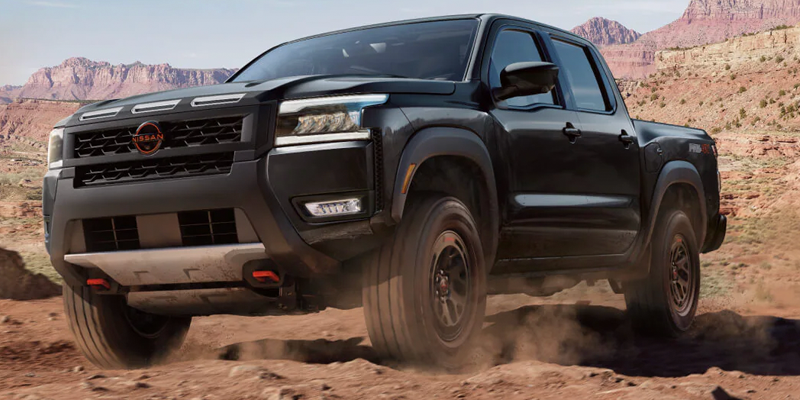 A black Nissan Frontier driving off-road through a dusty desert landscape with red rock canyons in the background.