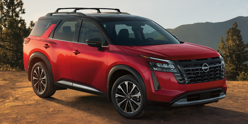A red Nissan Pathfinder with a black roof and roof rails parked in a mountainous outdoor setting.