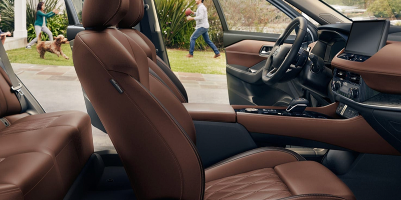 Interior view of a vehicle featuring brown leather seats with quilted stitching, with the front door open revealing a family playing in the yard in the background.