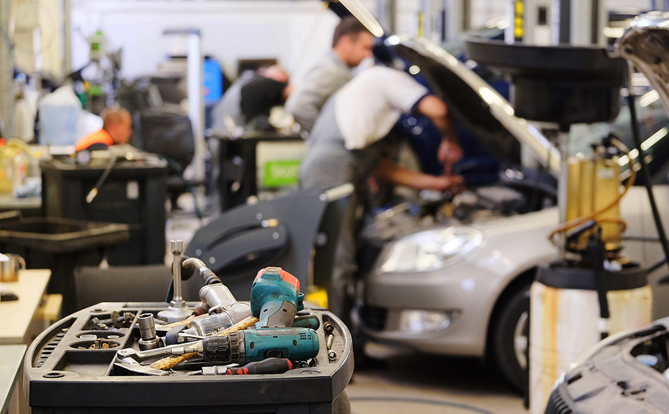 Close-up of a tool cart with drills and wrenches in a busy car repair shop, with mechanics working on vehicles in the blurred background.