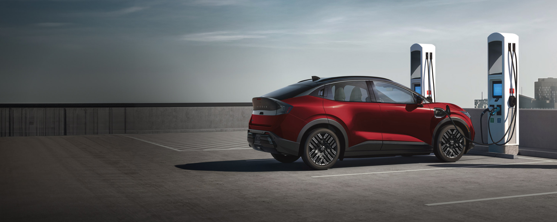 A red crossover SUV charges at an electric vehicle charging station with two tall white chargers under a bright sky.