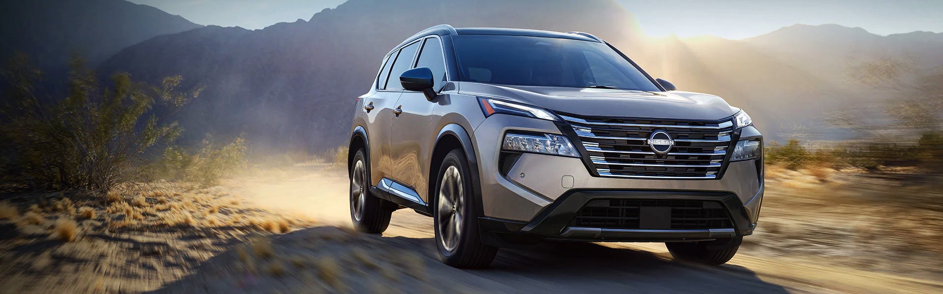 A pre-owned silver Nissan Rogue SUV driving along a dusty desert trail in front of mountains at sunrise.